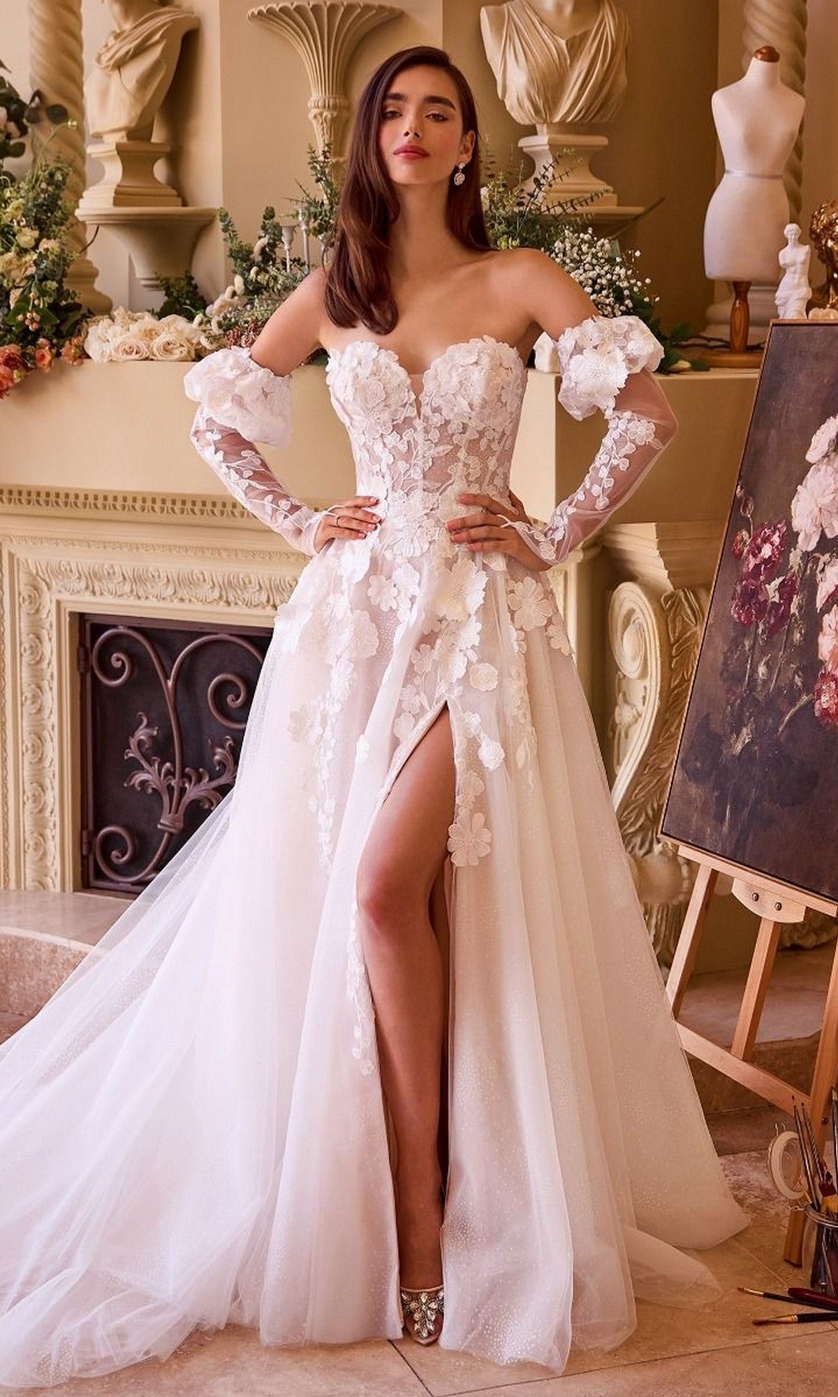 A woman in the Andrea and Leo WL017 off-shoulder long wedding dress with 3D floral appliques and a high slit stands indoors by a fireplace and an easel displaying a floral painting.